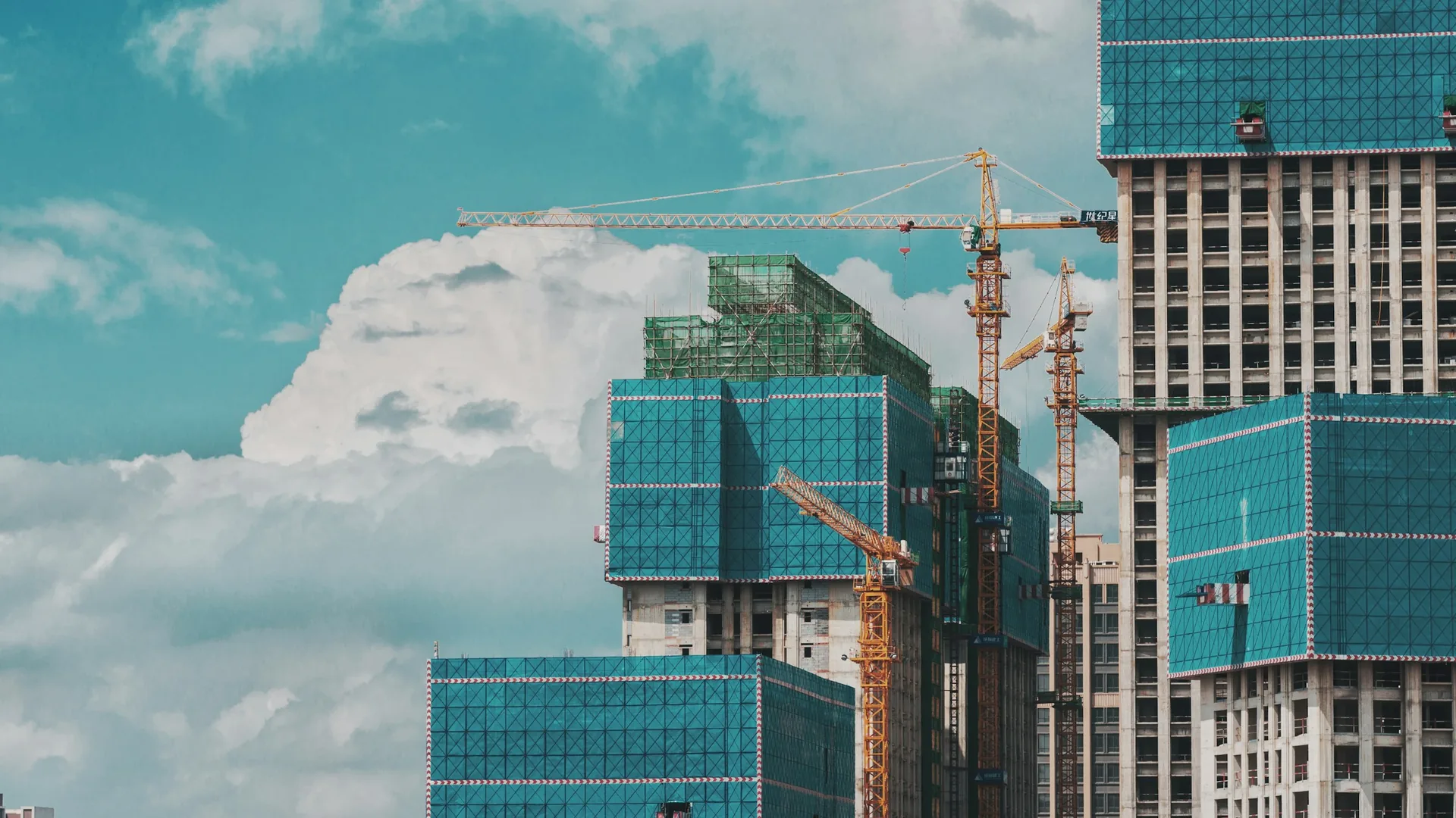 Yellow tower cranes beside high-rise buildings under construction, wrapped in blue and green protective screens.